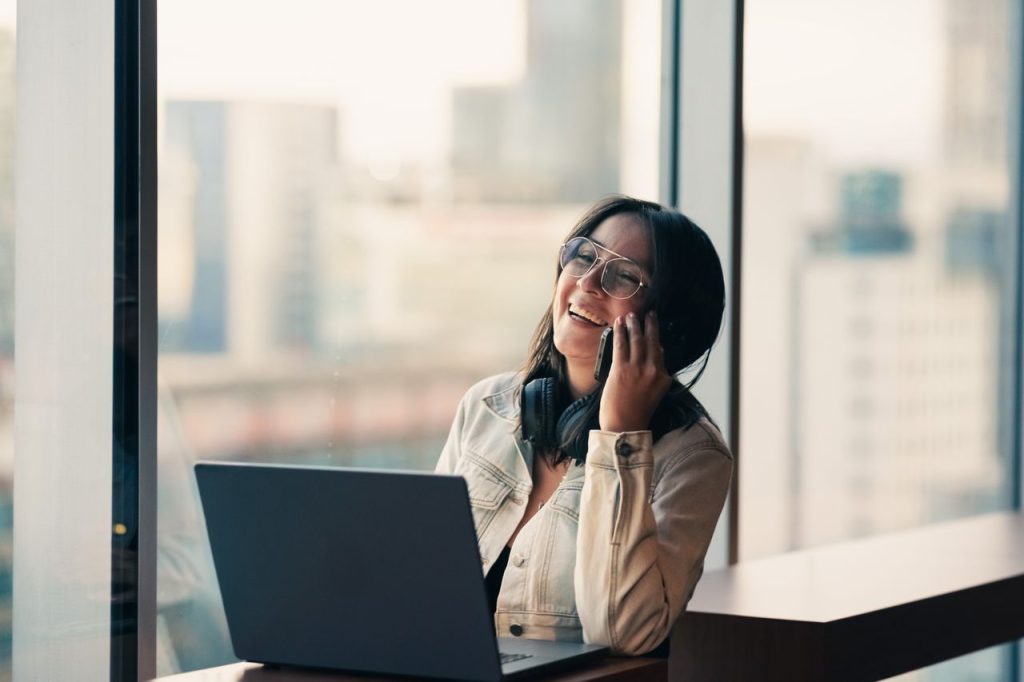 A joyful woman with glasses and headphones chats on phone by laptop in a modern office.