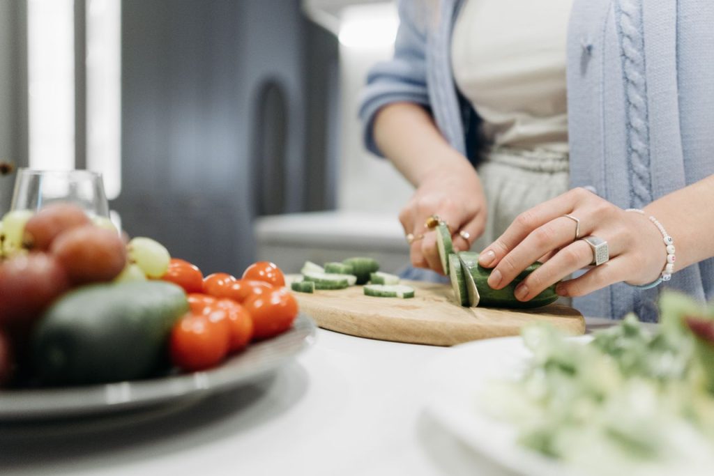 Close-up of a woman slicing cucumbers on a wooden board surrounded by fresh vegetables.